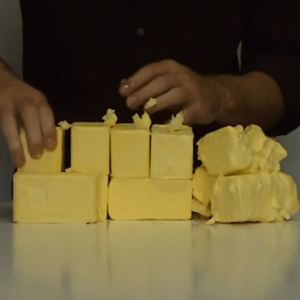 Yellow blocks of butter are stacked and formed on a table by the artist who stands behind it